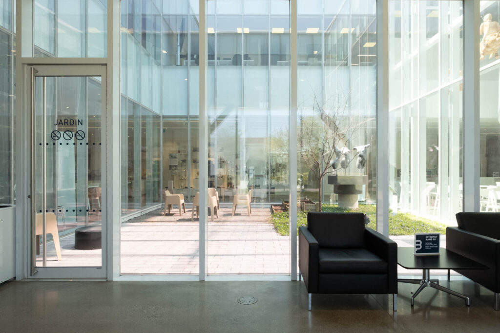 Interior of a contemporary library, showing a glass-enclosed patio in which are a set of chairs, planters and a sculpture representing three partial female torsos. The word "Jardin" is written on the door leading to the patio.