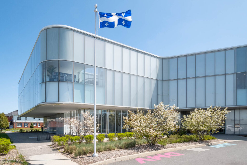 Exterior of a contemporary library, a three-story structure with gently curving glass walls. The ground floor is recessed from the upper stories, providing a covered area. In the foreground are flowering shrubs, a Quebec flag flying on a mast and a parking lot on which the letters DMV (for Drummondville) are painted.