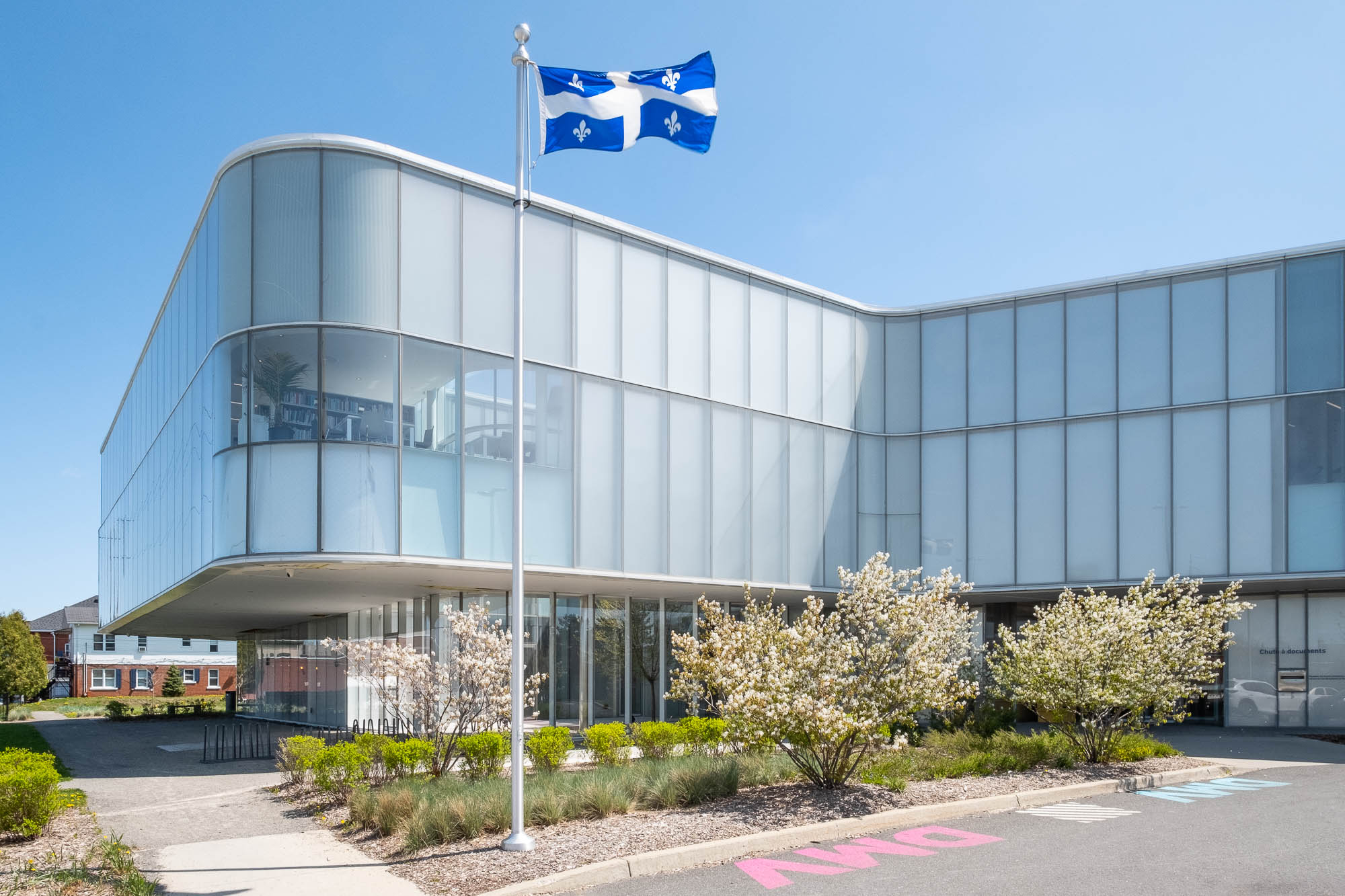 Exterior of a contemporary library, a three-story structure with gently curving glass walls. The ground floor is recessed from the upper stories, providing a covered area. In the foreground are flowering shrubs, a Quebec flag flying on a mast and a parking lot on which the letters DMV (for Drummondville) are painted.