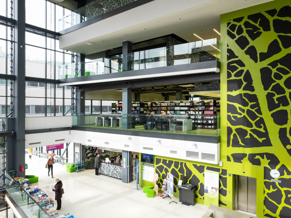 Interior of a contemporary library, showing the library's entrance and welcome desk in a large glass atrium. Three floors of bookshelves and worktables can be seen above the service desk. The elevator shaft is decorated with a leaf pattern in green and black.