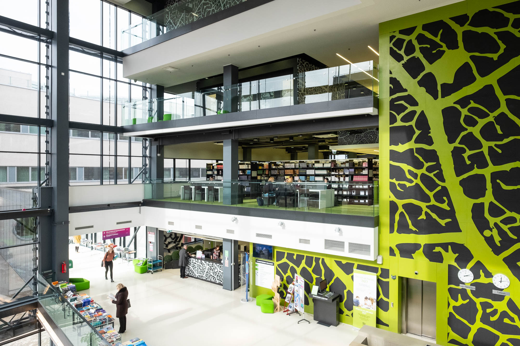 Interior of a contemporary library, showing the library's entrance and welcome desk in a large glass atrium. Three floors of bookshelves and worktables can be seen above the service desk. The elevator shaft is decorated with a leaf pattern in green and black.