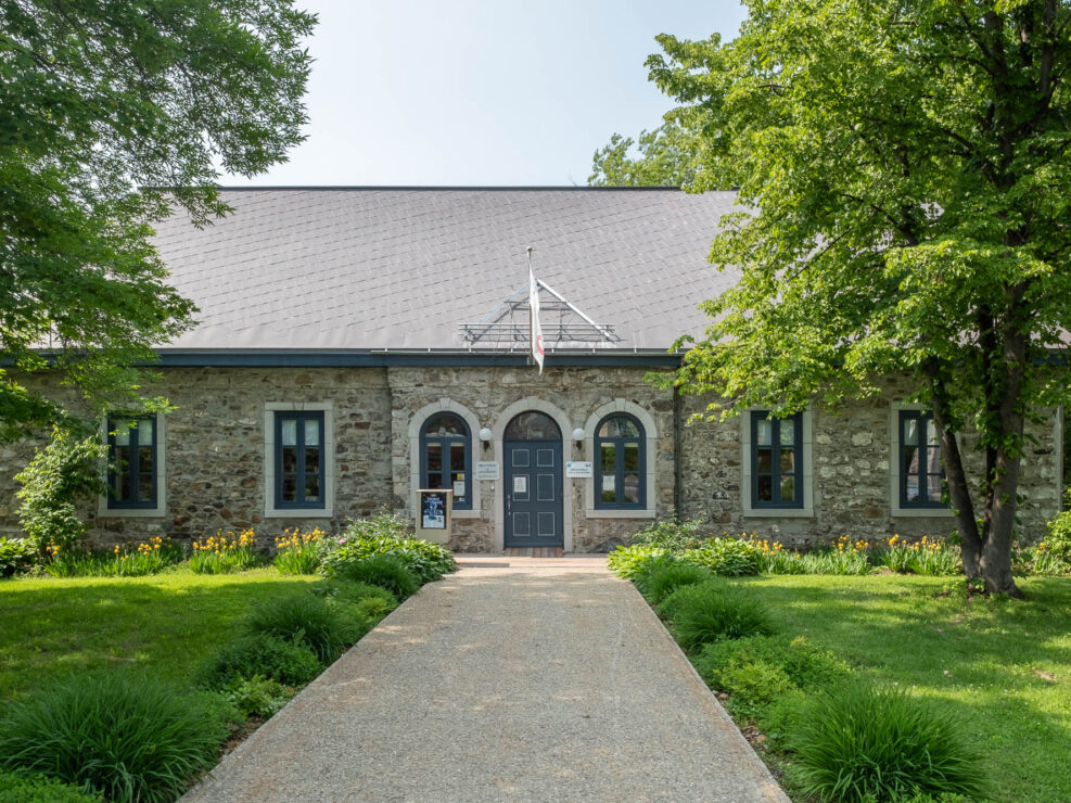 Exterior of a presbytery converted to a library. It is a low, single story stone building with simple pitched roof. It is surrounded by mature trees and a well-kept lawn.