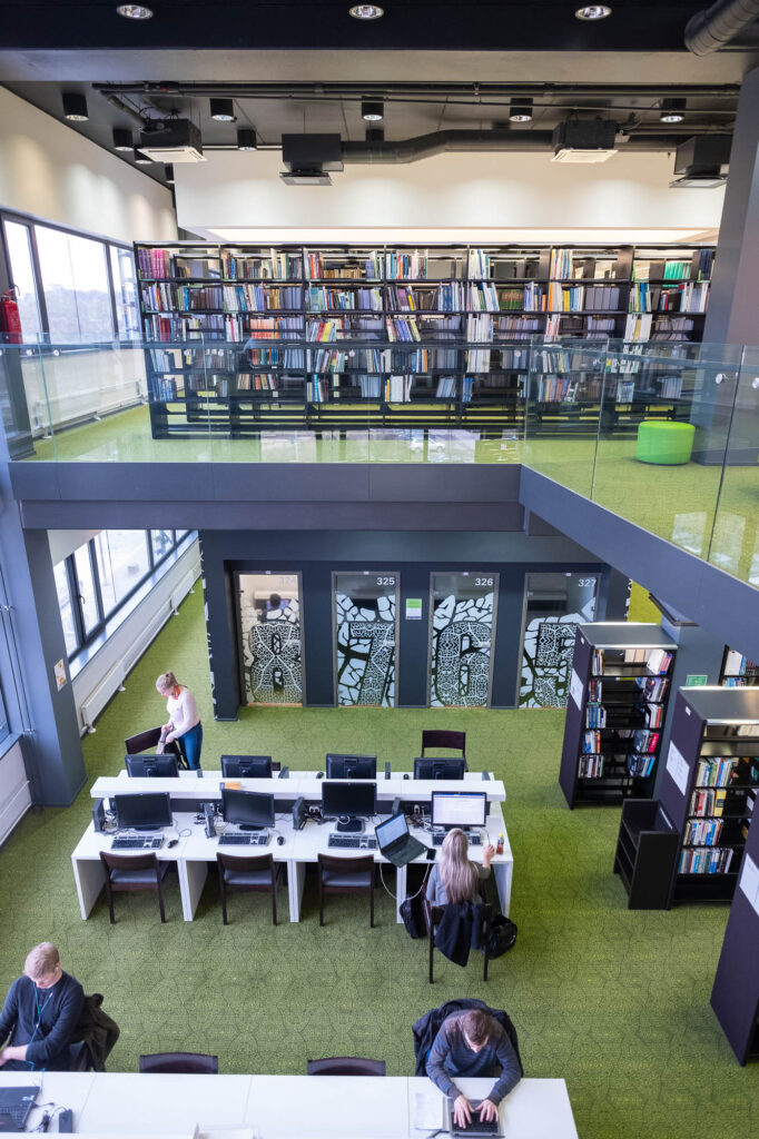 Interior of a contemporary library, looking down from a mezzanine into a level with bookshelves, work tables on which students are working at computers, and individual study rooms.