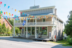 Exterior of a "boomtown"-style general store converted to a library. It is a rectangular building covered in wood planks with a gallery in front supported by timber posts and crowned by a pastiche facade. The number 1917 is written on top. Colourful bunting and Quebec flags hang across the street in the front.