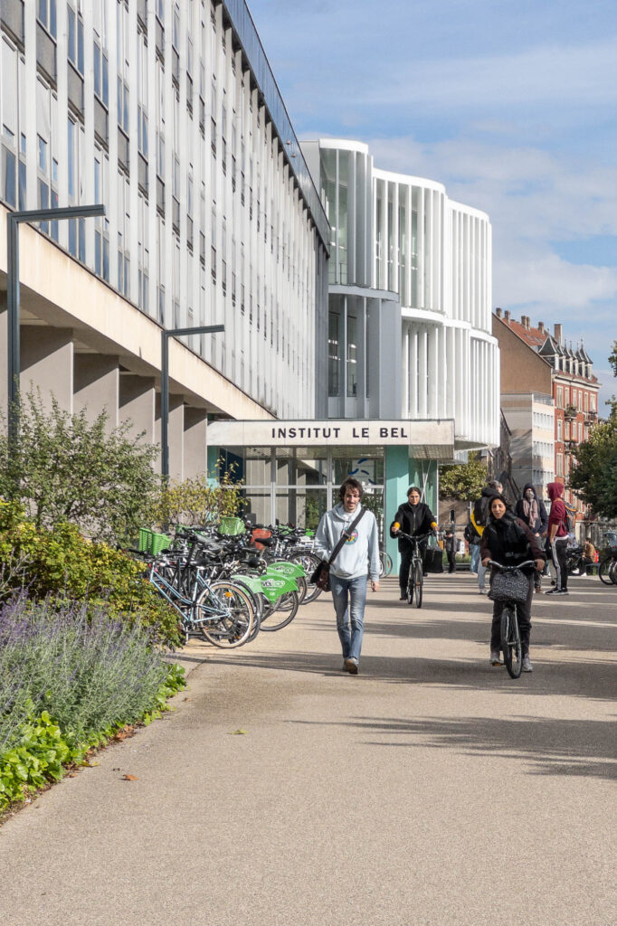 Campus street scene with students walking and biking. To the left is a Modernist building with an awning inscribed with the words "Institut Le Bel". In the background is a contemporary curvy building with vertical brise-soleils.