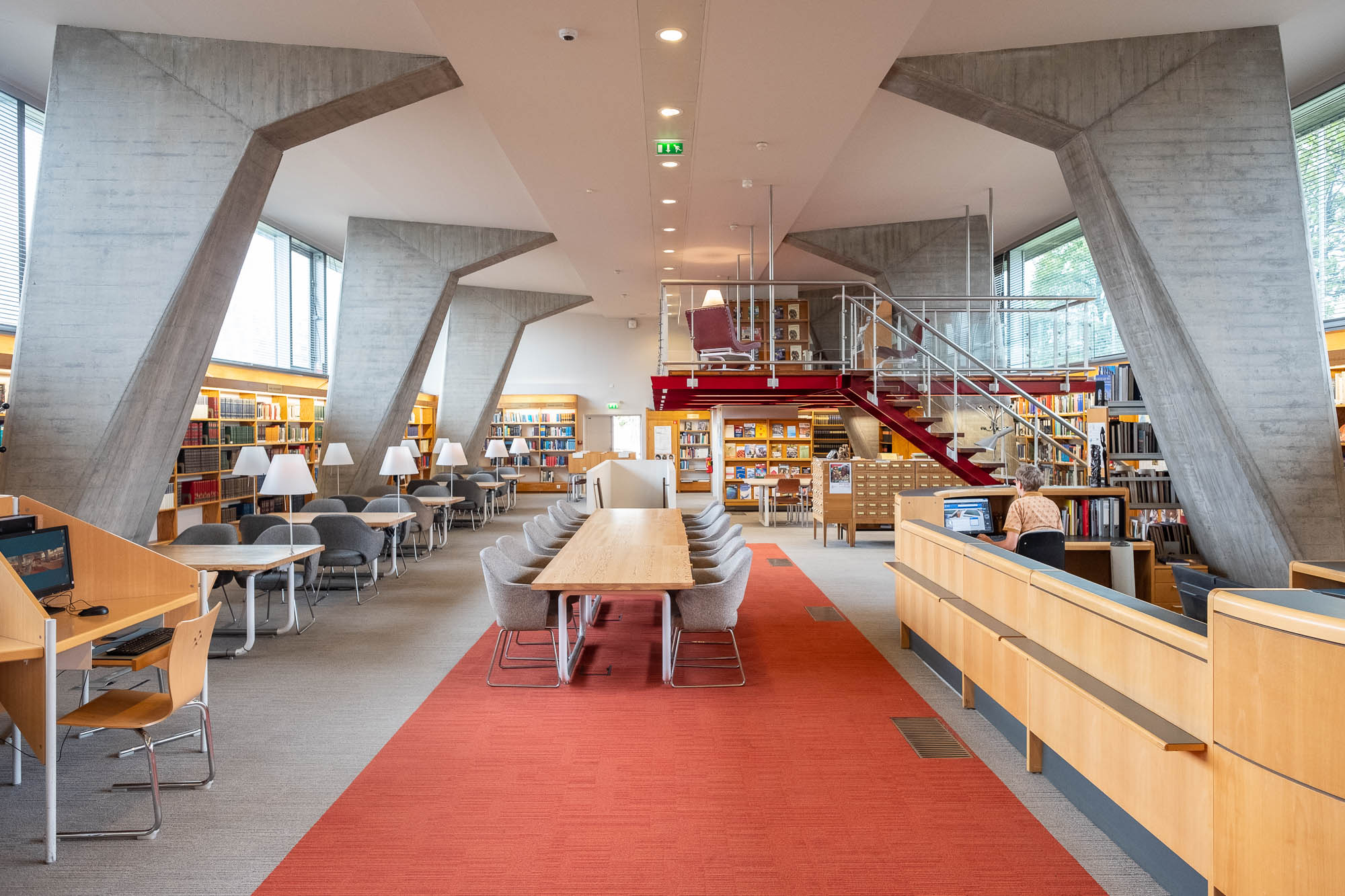 Interior of a Modernist library, a large double-height space supported by large, geometric concrete columns. At the right is the welcome desk at which a person is working at a computer, facing away from the camera.