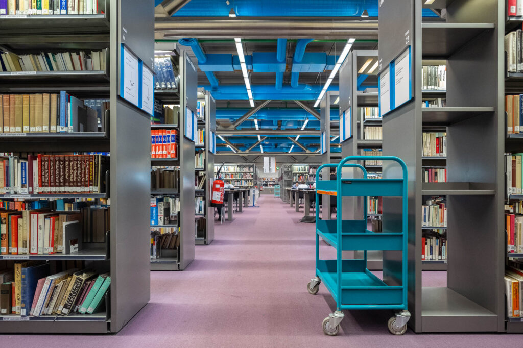 A library interior with tall bookshelves filled with books, a turquoise book cart with empty shelves, and rows of shelves extending into the distance under a ceiling with exposed blue pipes.