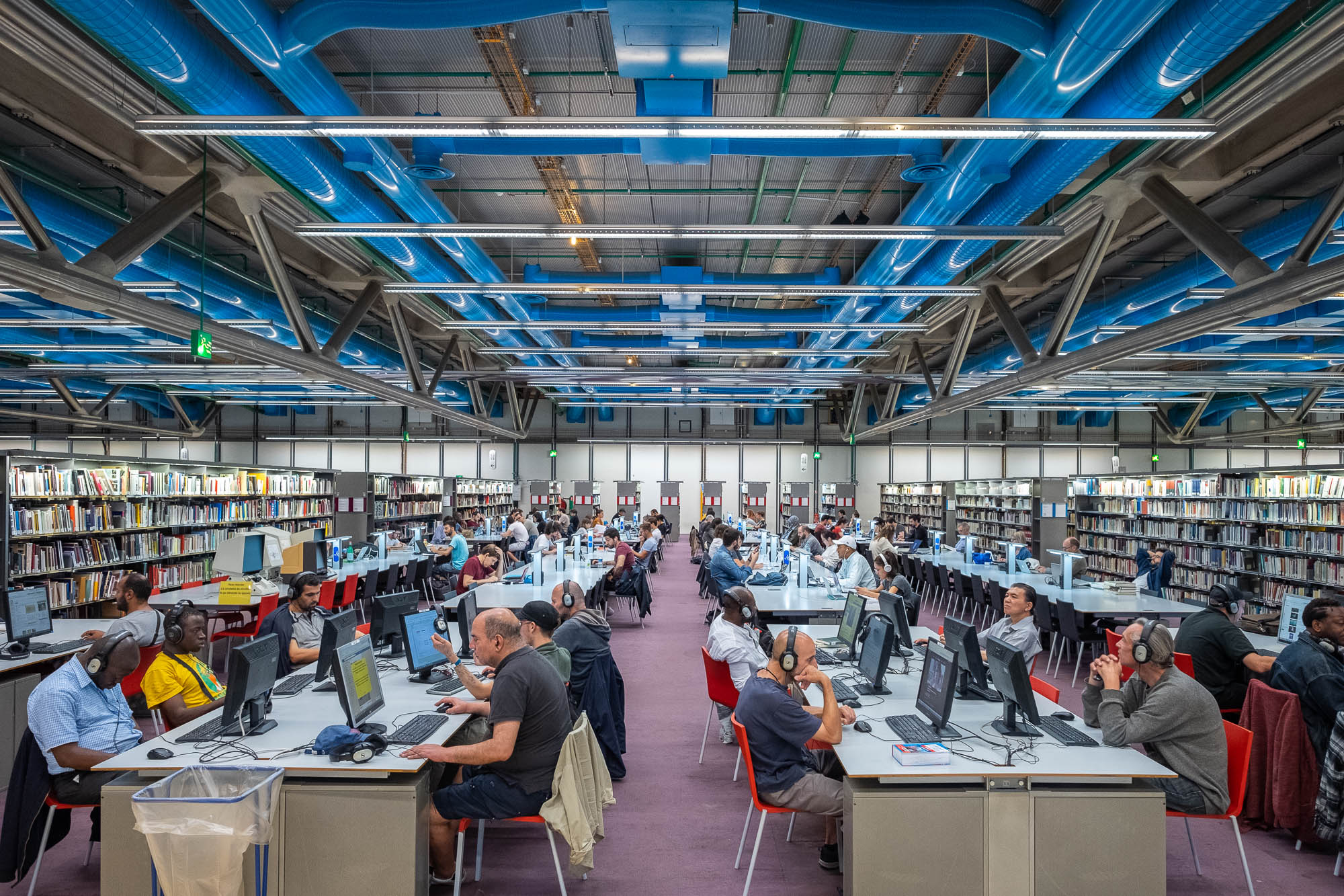 Large, modern library with rows of tables where many people are seated, studying or using laptops. Bookshelves are visible in the background. The ceiling has visible blue ducts and industrial lighting.