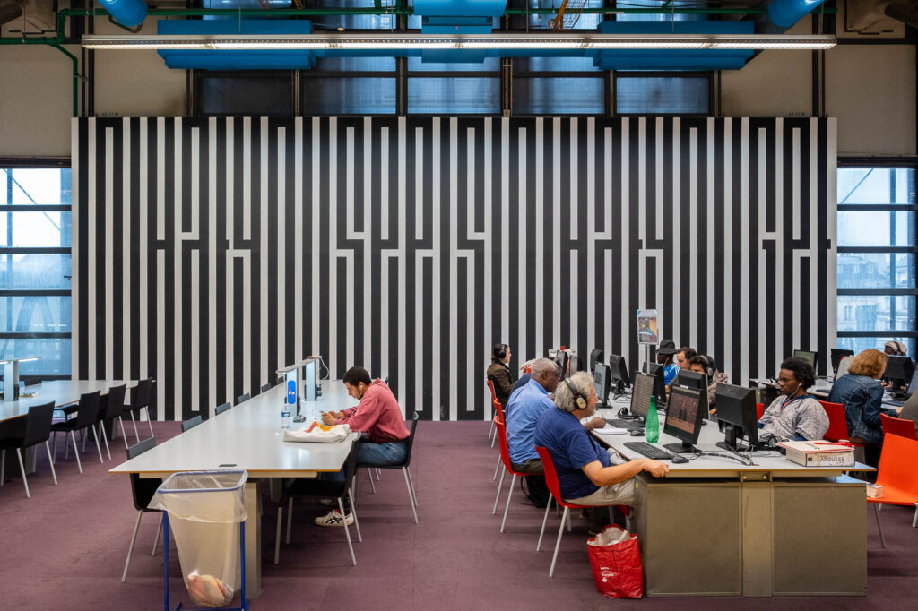 People are seated at desks in a modern library, working on computers. The room features a large wall with a black and white geometric striped pattern and large windows on either side.