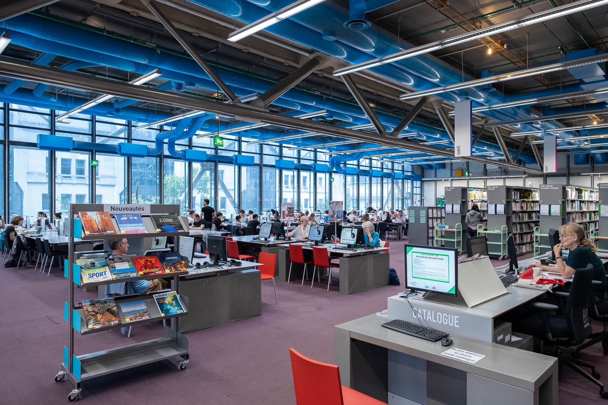 A spacious modern library with large windows, several rows of desks with people working on computers, bookshelves, and a display stand of books in the front.