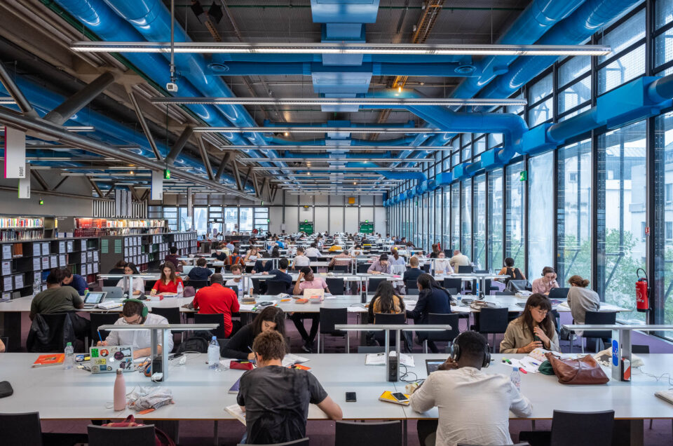 Large, modern library with rows of tables where many people are seated, studying or using laptops. Bookshelves are visible in the background. The ceiling has visible blue ducts and industrial lighting.