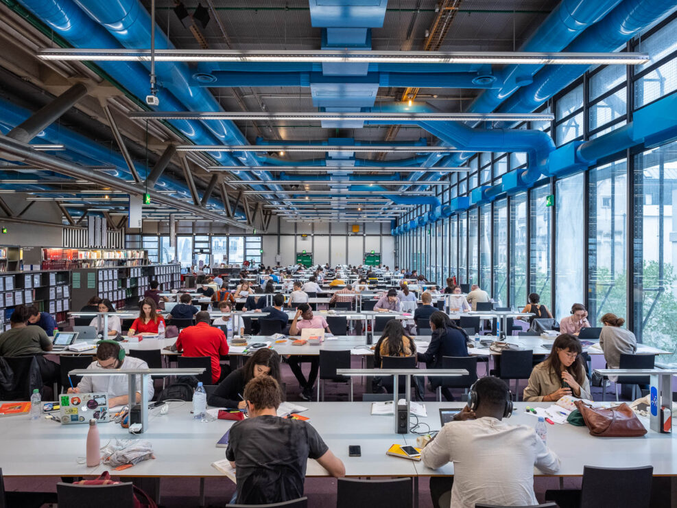 Large, modern library with rows of tables where many people are seated, studying or using laptops. Bookshelves are visible in the background. The ceiling has visible blue ducts and industrial lighting.