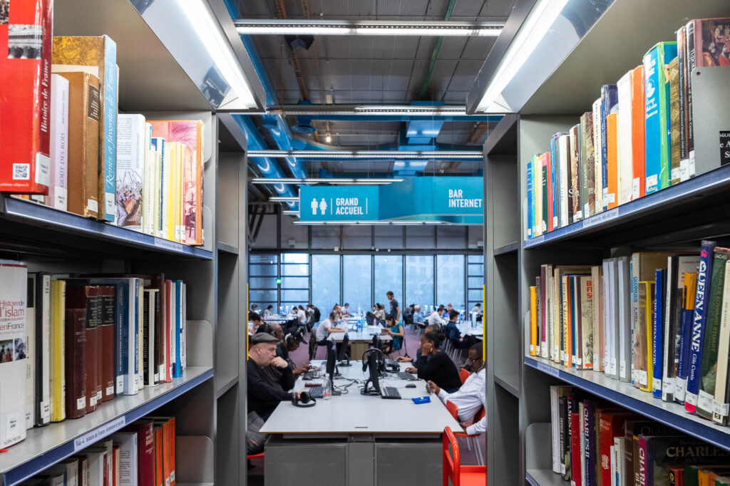 Bookshelves line both sides in the foreground, framing a view of a library study area where people sit at tables using computers. Overhead signs read Grand Accueil and Bar Internet.