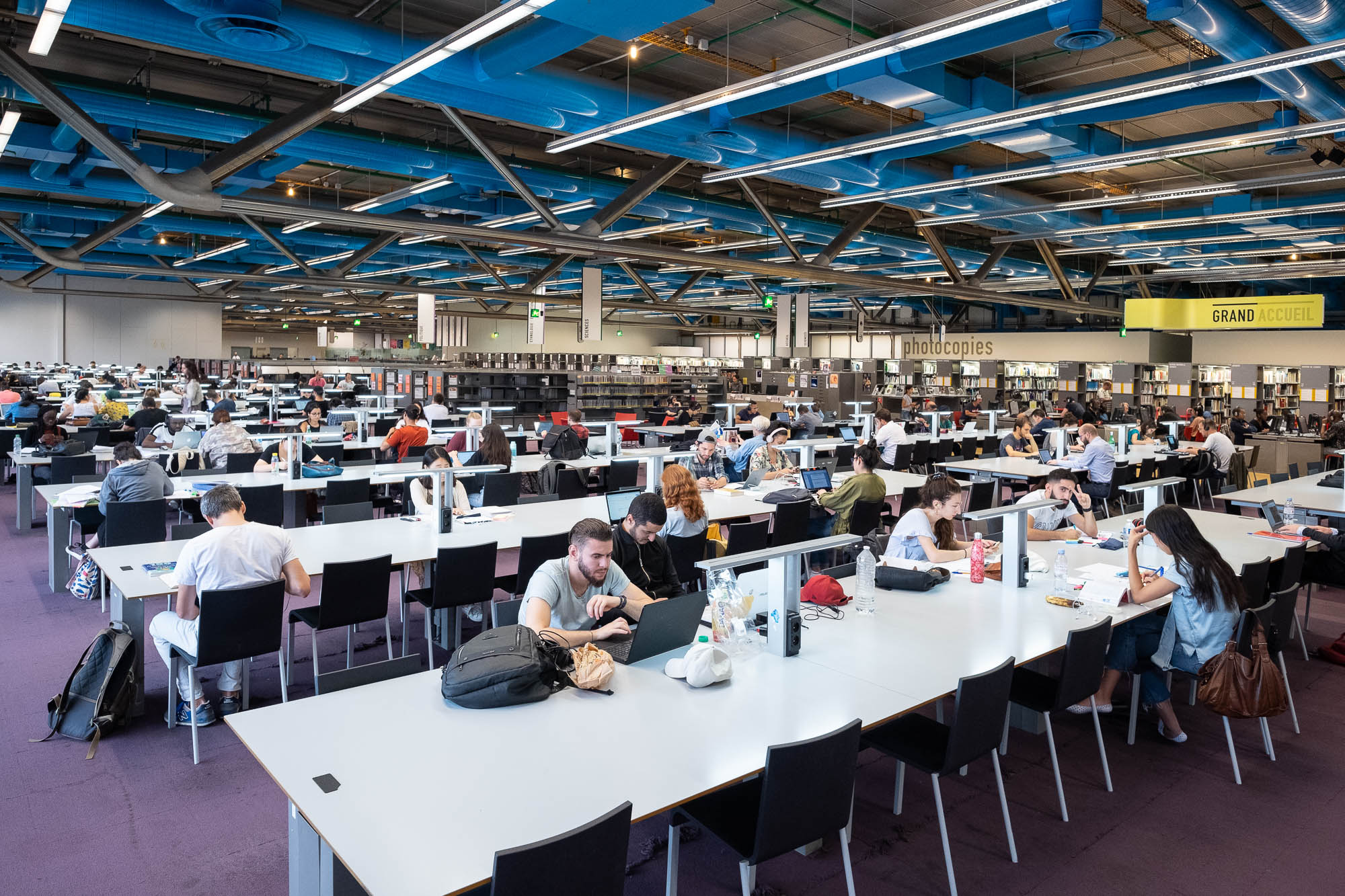 Large, modern library with rows of tables where many people are seated, studying or using laptops. Bookshelves and photocopy stations are visible in the background. The ceiling has visible blue ducts and industrial lighting.