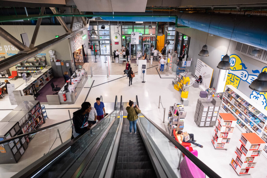 People use an escalator inside a modern library with bookshelves, seating areas, colorful decor, and individuals browsing, reading, or walking. Bright lighting and open design are visible.
