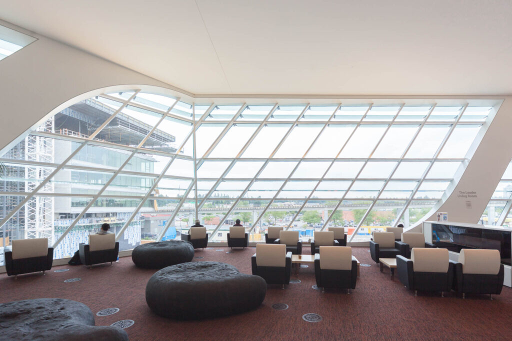 A contemporary lounge area with large slanted windows, black and white armchairs, circular black ottomans, and a view of the runway and tarmac outside. A few people are seated, and natural light fills the space.