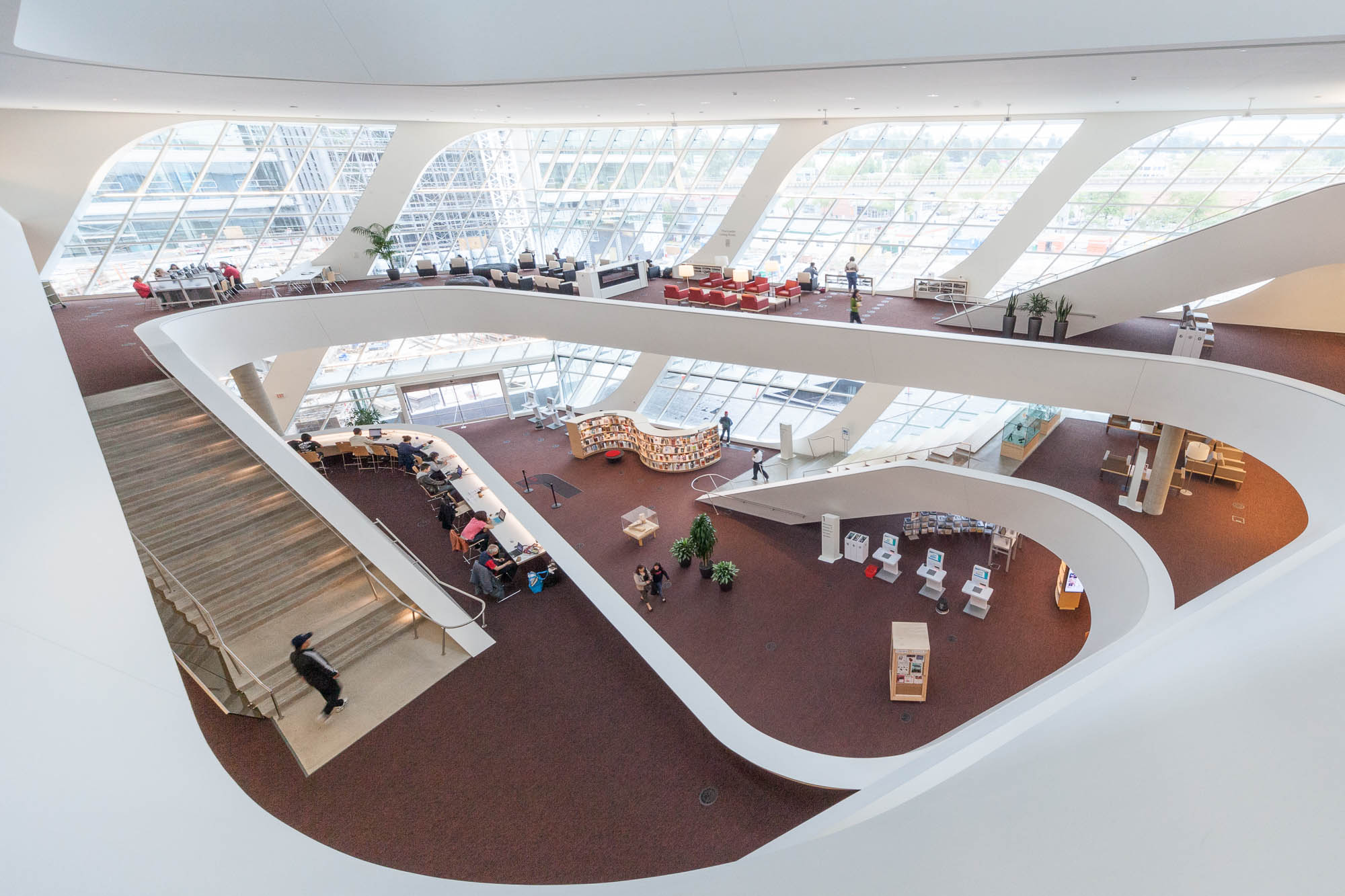 Interior of a contemporary library with curved white balconies, large windows allowing natural light, multiple floors with bookshelves, and people reading or working at tables.