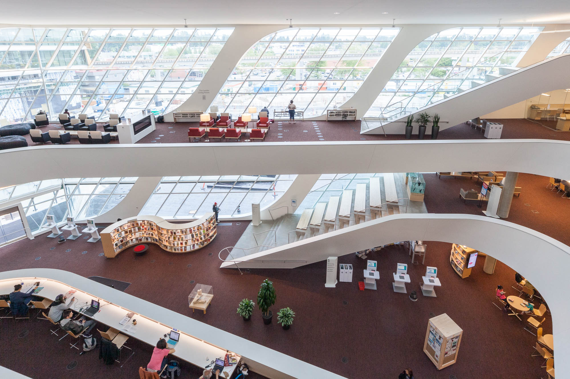 Interior of a contemporary library with curved white balconies, large windows allowing natural light, multiple floors with bookshelves, and people reading or working at tables.