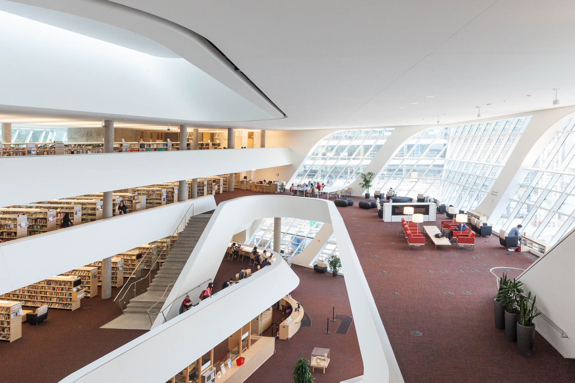Interior of a contemporary library with curved white balconies, large windows allowing natural light, multiple floors with bookshelves, and people reading or working at tables on lower levels.