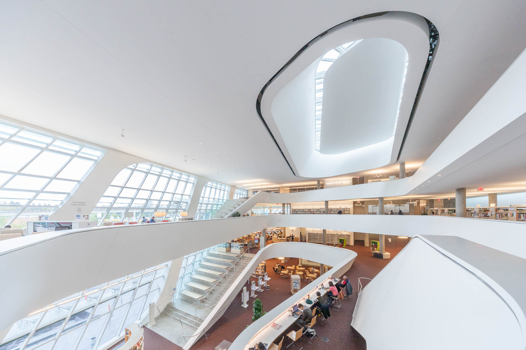 Interior of a contemporary library with curved white balconies, large windows allowing natural light, multiple floors with bookshelves, and people reading or working at tables on the lower level.