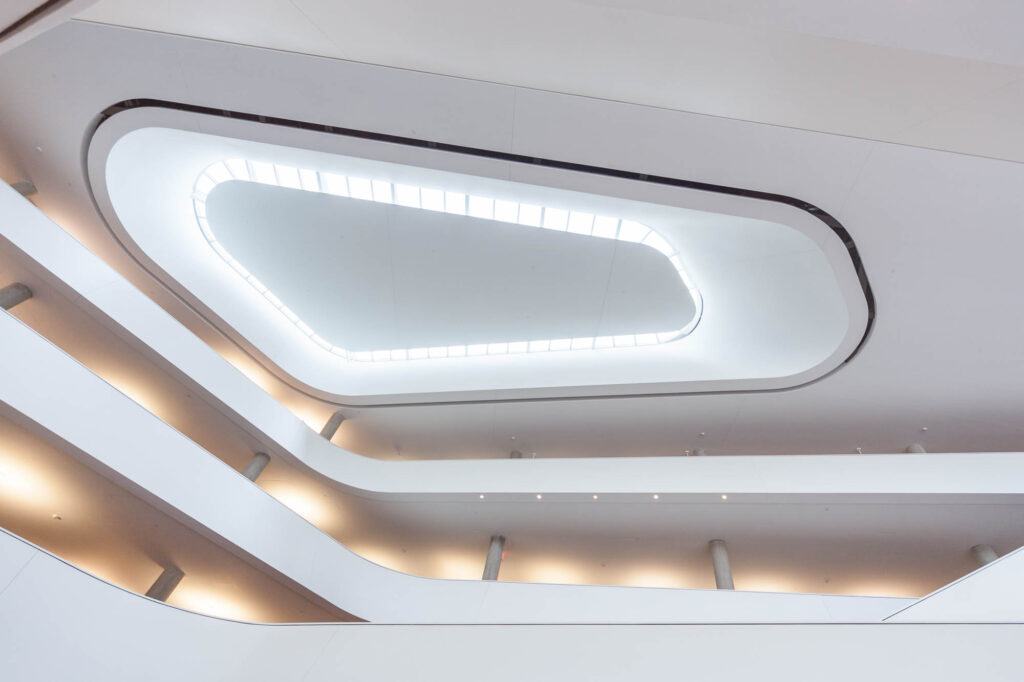 Interior view of a contemporary building with white walls, curved balconies, and a large oval-shaped skylight in the ceiling, allowing natural light to fill the space.