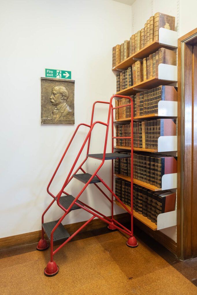 Interior of a library, showing wall shelves filled with old books and a stepladder. On the wall is a portrait of a man with the words "Master AWC 1911 WR Lethaby". A fire exit sign sits immediately above the portrait.
