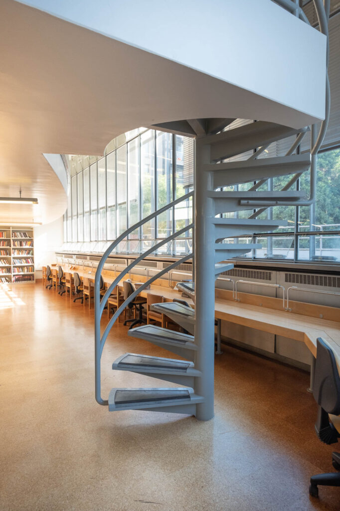Interior of a modernist library showing a spiral staircase. Behind it is a large window with a long wooden table.