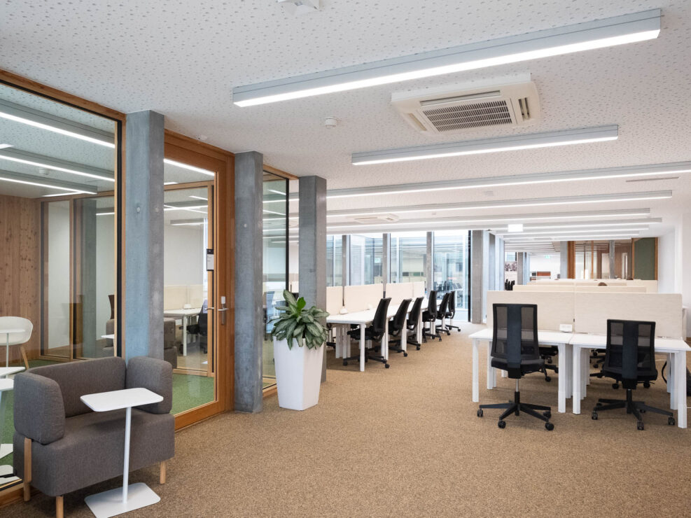 Interior of a contemporary library with rows of tables and computer chairs, and a comfy chair at the front.
