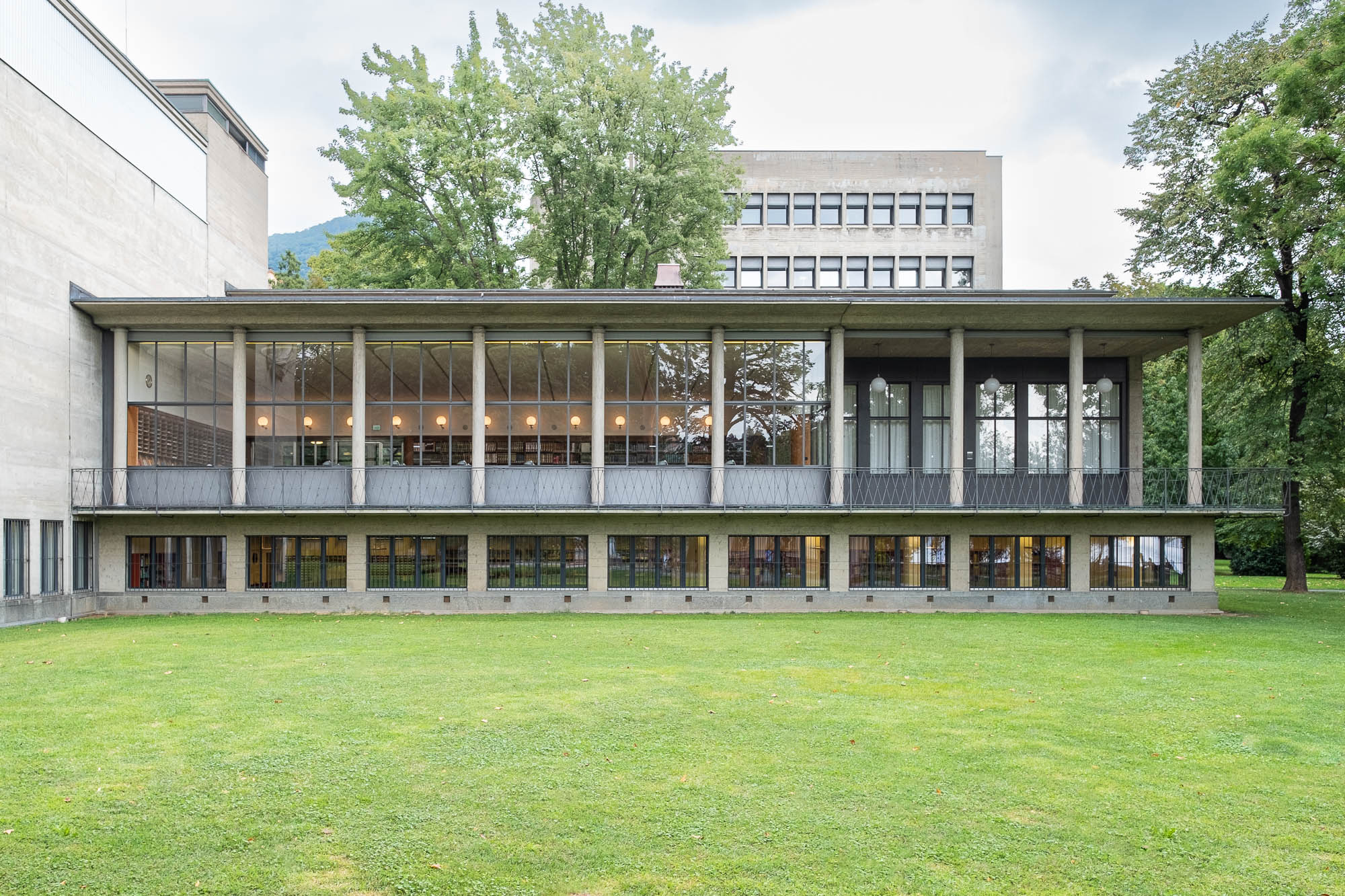 Exterior of a modernist library in a park surrounded by trees. It is an L-shaped building. The wing facing the image is fronted by a colonnade, two third of which are filled with glazing through which a reading room can be seen. The last third is open to a terrace.
