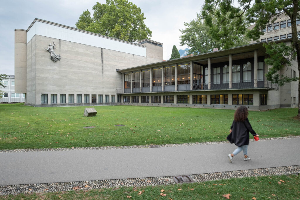 Exterior of a modernist library in a park surrounded by trees. It is an L-shaped building. The wing to the left has a largely blind concrete facade, bearing a sculpture representing a draped woman with her arms raised (Minerva). The wing to the left is open by glazing and fronted by columns. A person is walking on a path in front of the image.