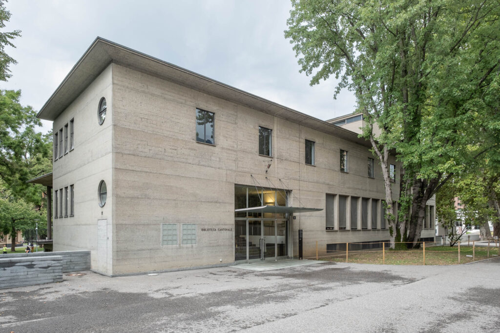Exterior of a modernist library. It is a rectangular concrete building with rectangular windows and two round windows. The entrance is square and covered with a thin metallic porte cochère. The words "Biblioteca Cantonale" are written left of the entrance.