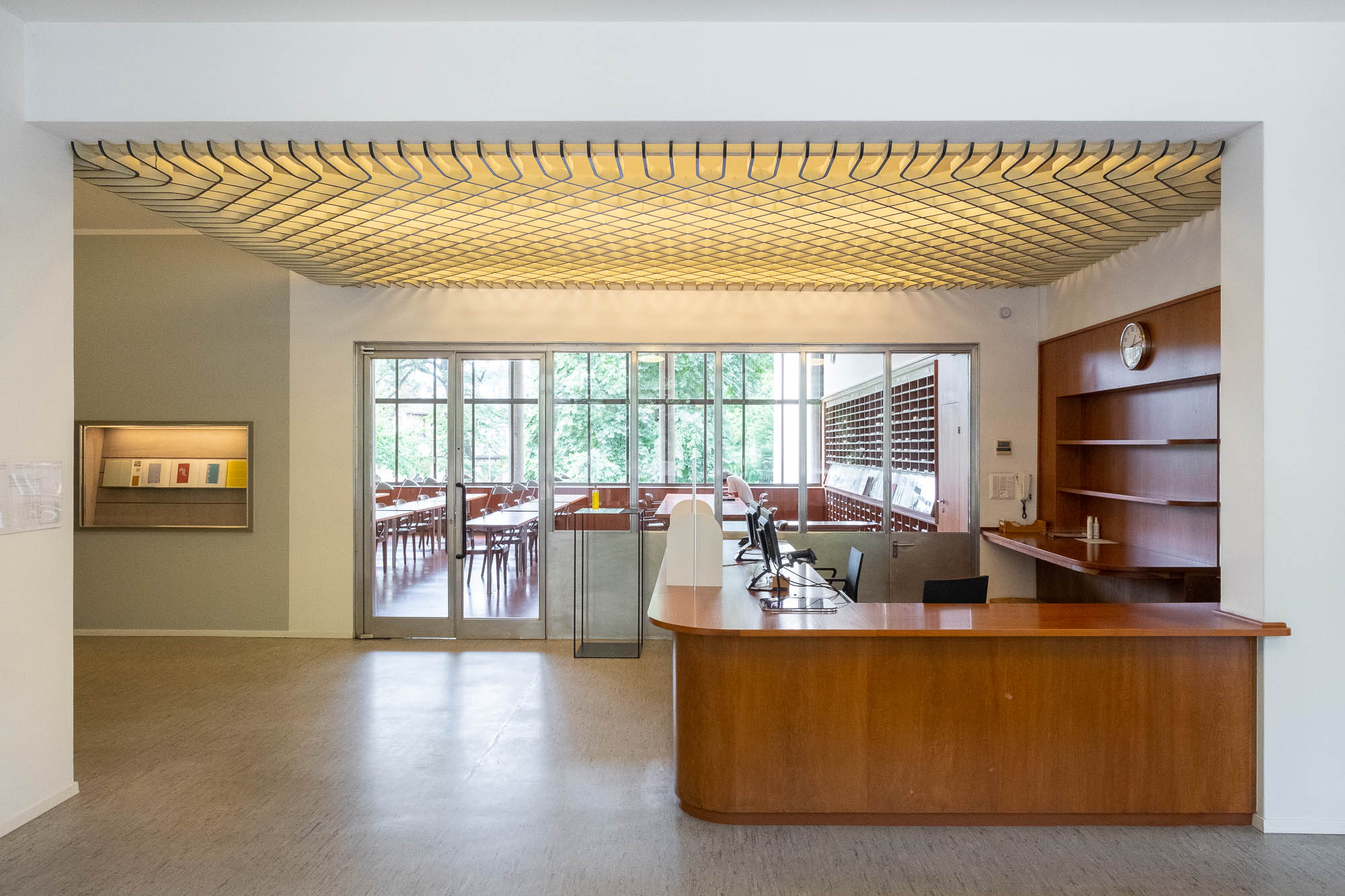 Interior of a modernist library, showing a wooden service desk with rounded edges. Behind it we can see a reading room through glazing.