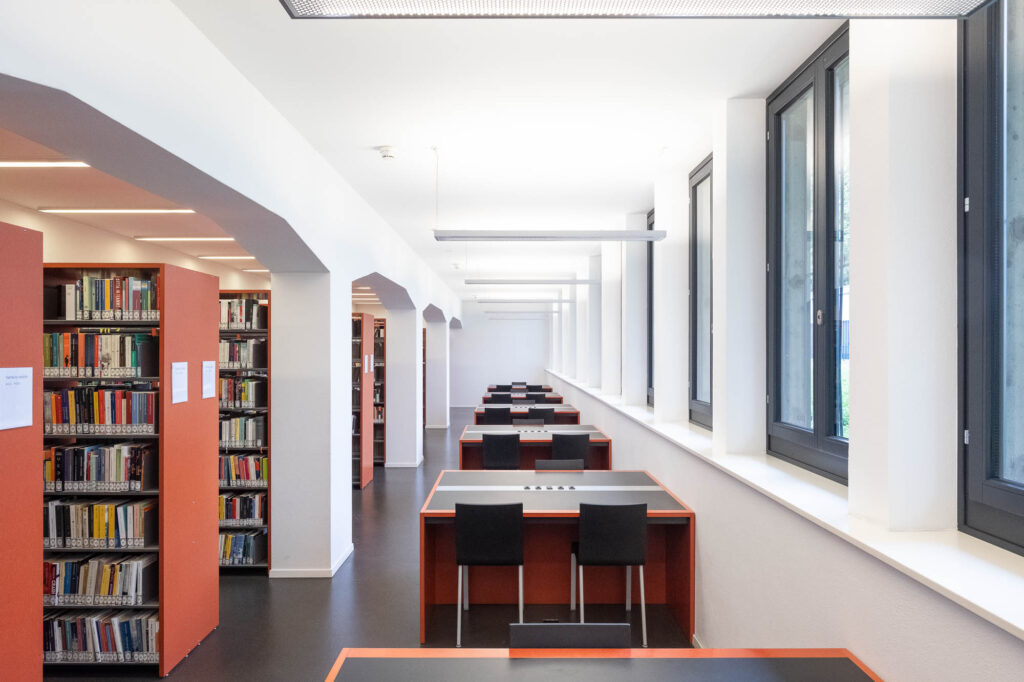 Interior of a library with a row of small work cubicles next to rectangular windows. To the left are bookshelves under broken arches.