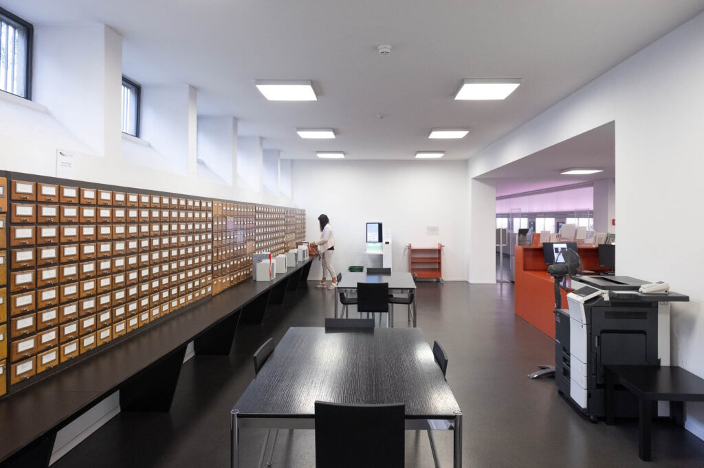 Interior of a library with a card catalogue to the left and a person utilizing a checkout machine at the back.