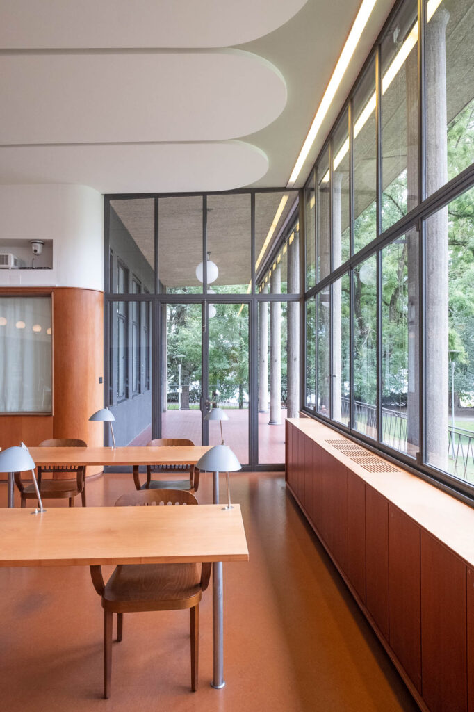 Interior of a modernist library, with large windows at the back and the left. Through them we see columns and trees.