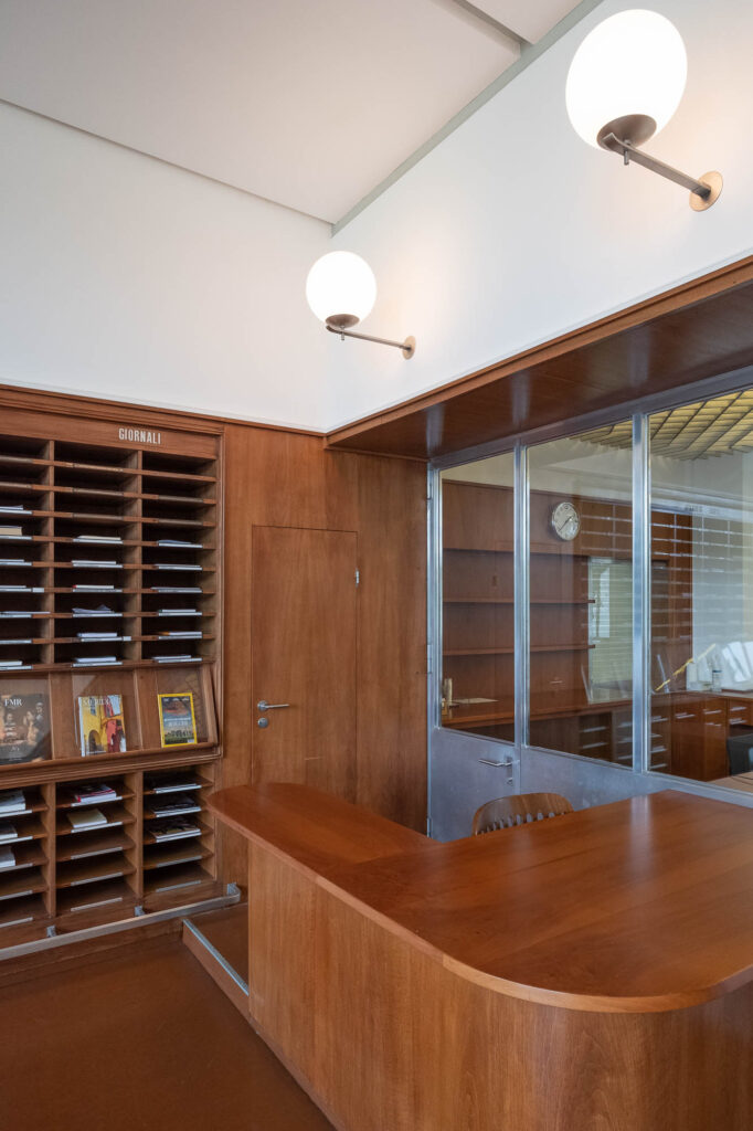 Interior of a modernist library, showing a small wooden service desk with rounded corners. Behind it is a wall of deep shelves and angled magazine display, topped with the word "Giornali". Globe lights are set on the wall to the right.