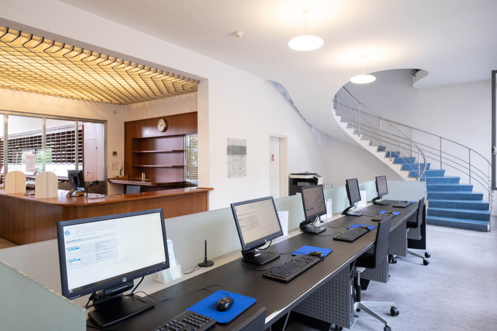 Interior of a modernist library. To the left there is a wooden service counter, behind it are windows opening to a reading room. To the right is a spiral staircase raising to the floor above. There is a bank of computers in the front.