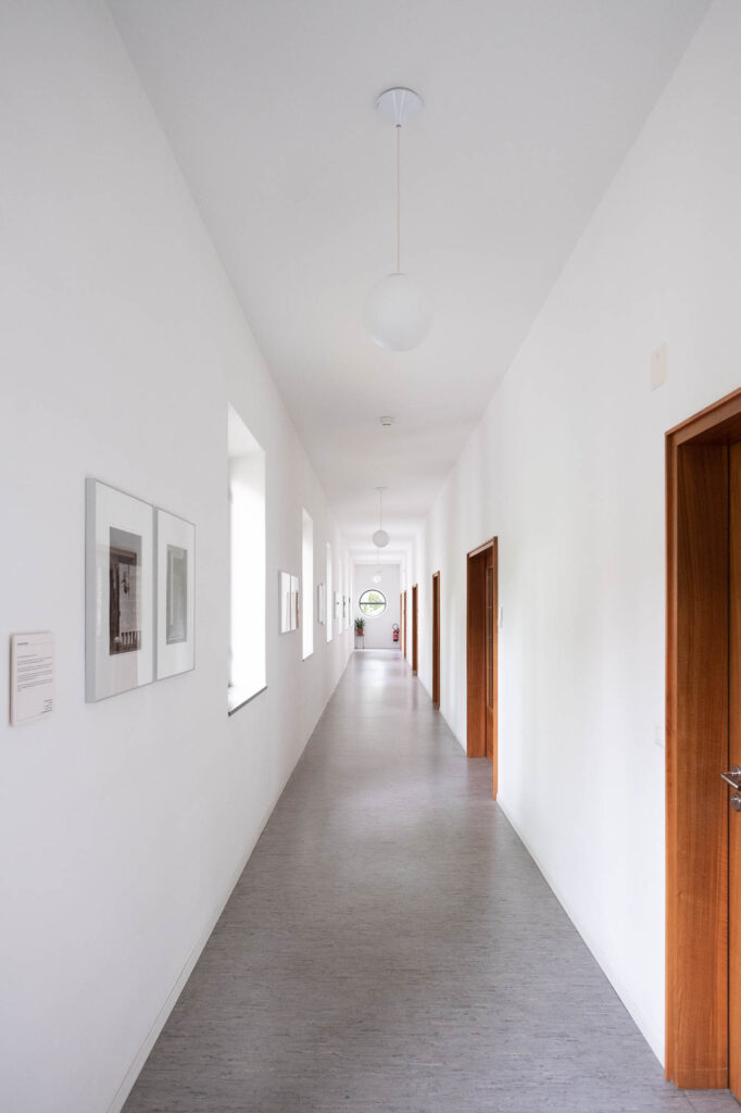 Interior of a modernist library, showing a long whitewashed corridor flanked by wooden door insets. There are globe lights on the ceiling. The end of the corridor opens with a round window.
