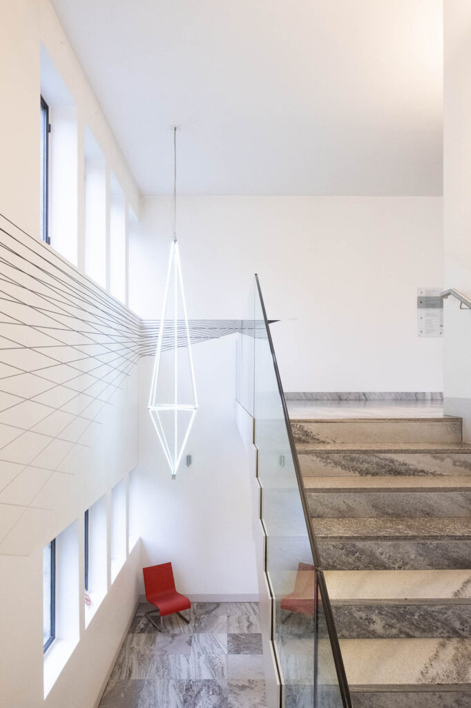 Interior of a modernist library, showing marble stairs surrounded by white walls. A contemporary geometrical chandelier hangs from the ceiling and geometric lines decorate the wall.