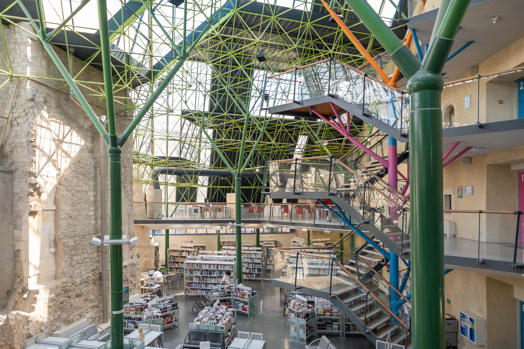 Interior of a library inside a converted former hospital, a high glass roof on metal stilts covers an area bound by original stone walls. The metal stilts branch out as they rise and are painted green, recalling tree trunks.