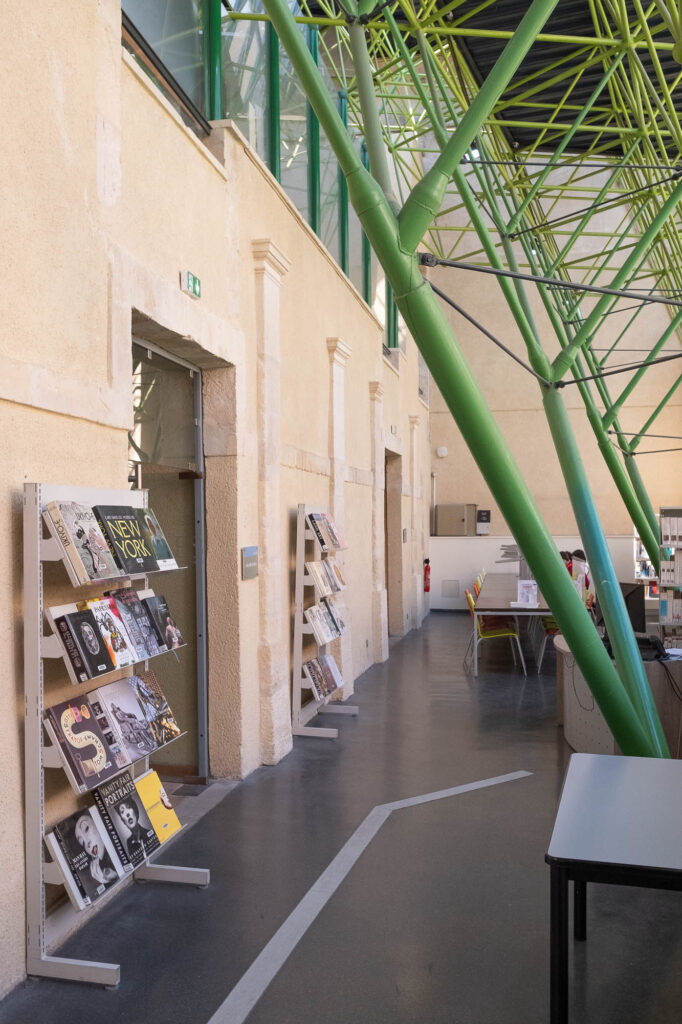 Interior of a library inside a converted former hospital, a high glass roof on metal stilts covers an area bound by original stone walls. The metal stilts branch out as they rise and are painted green, recalling tree trunks.