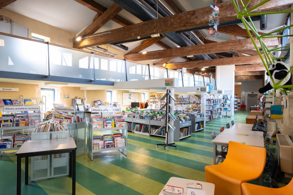 Interior of a library inside a converted former hospital, with visible timber beams. The area displayed is the children's section, with low bookshelves, colourful furniture and plush toys.