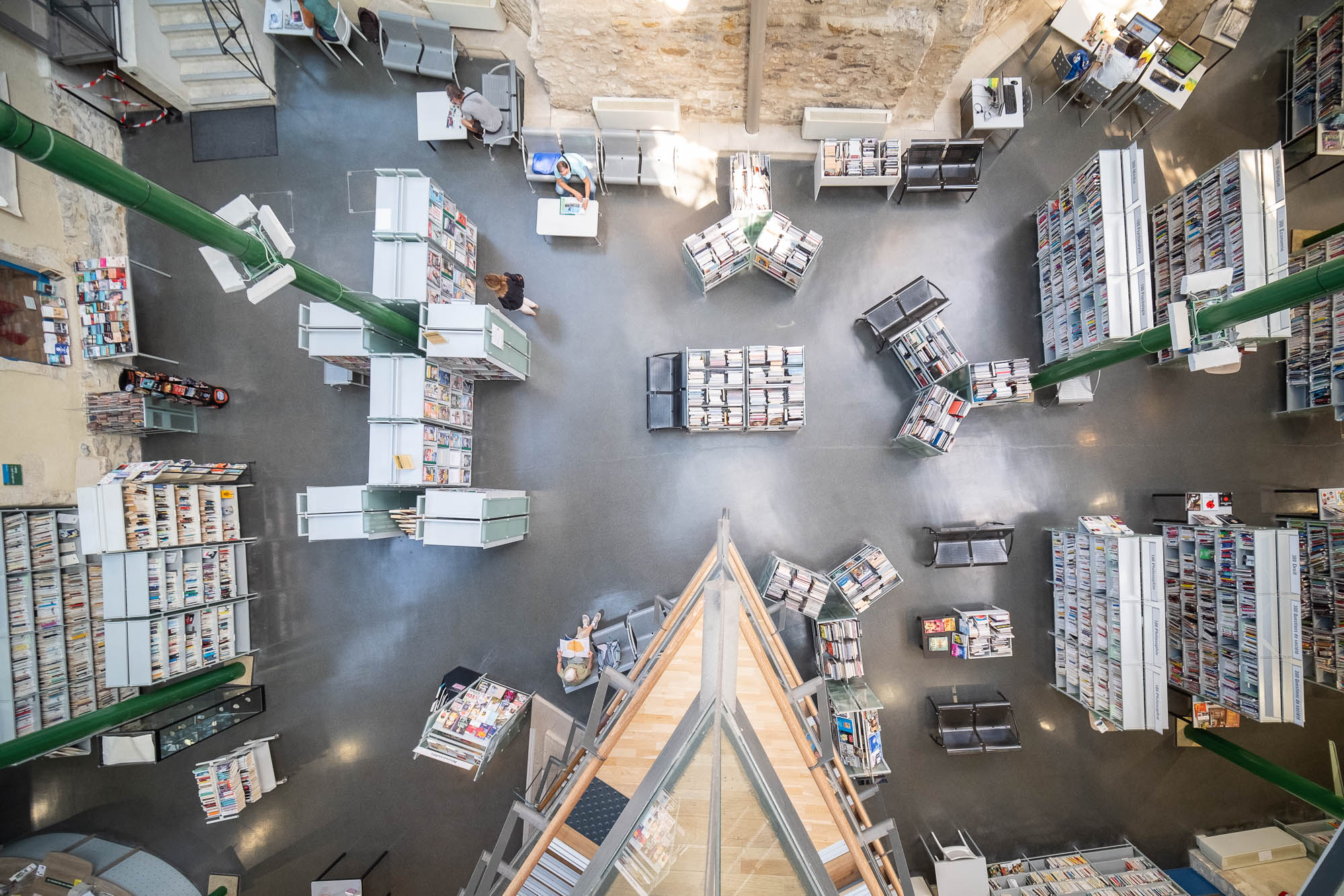 Interior of a library inside a converted former hospital, looking down from a pointed balcony towards the ground floor where rows of bookshelves alternate with seats and tables.
