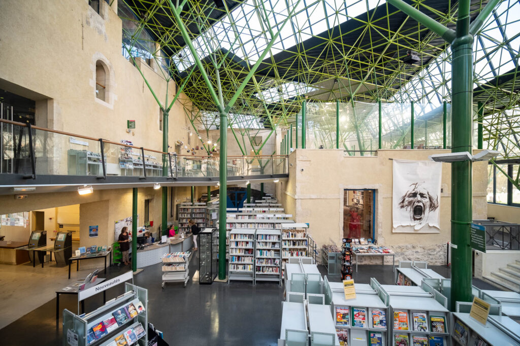 Interior of a library inside a converted former hospital, a high glass roof on metal stilts covers an area bound by original stone walls. The metal stilts branch out as they rise and are painted green, recalling tree trunks.