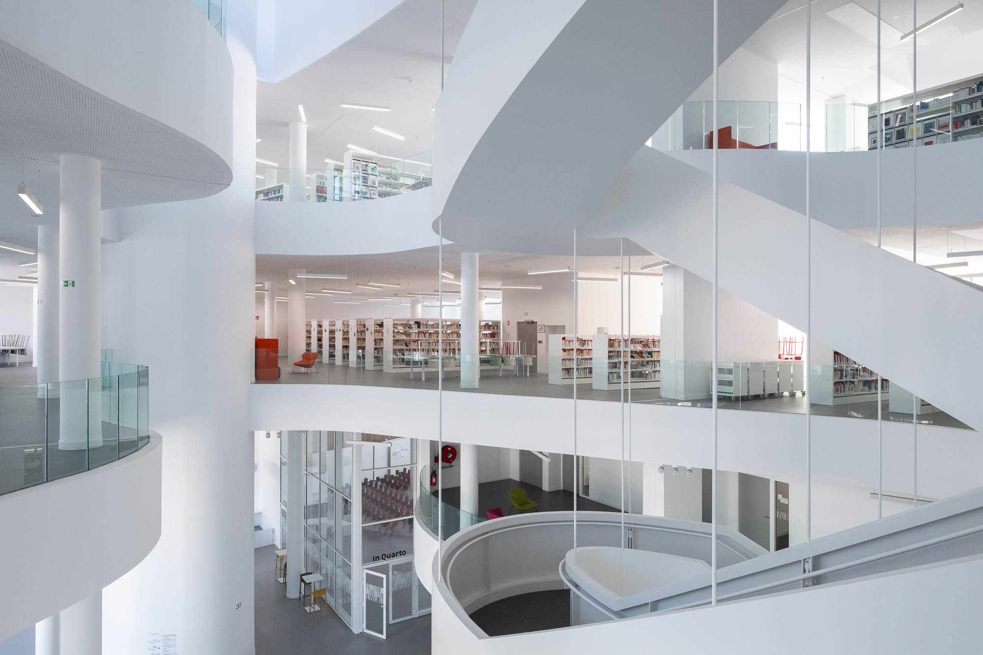 A contemporary, multilevel library with curved balconies, ramps and stairs. Filled bookshelves and colourful seats contrast against the grey and white surroundings.