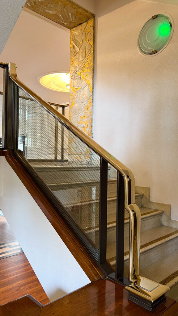 Staircase in an Art-Deco building, with gilded wall decorations on top and a golden handlebar. Up on the wall, a round light fixture glows faintly in green.