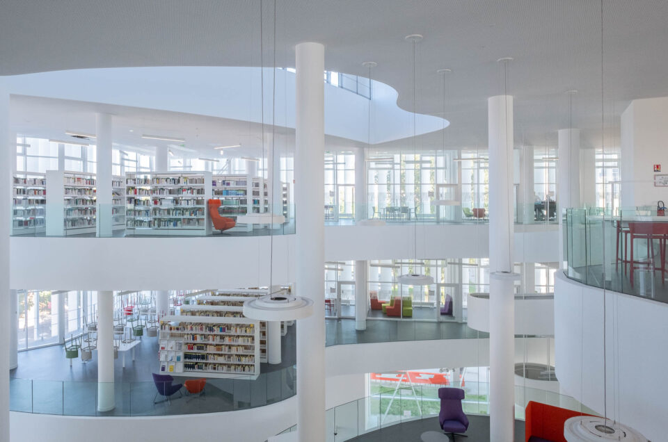 A contemporary, multilevel library with curved balconies and tall white columns. Filled bookshelves and colourful seats contrast against the grey and white surroundings. Natural light comes from large windows at the back.