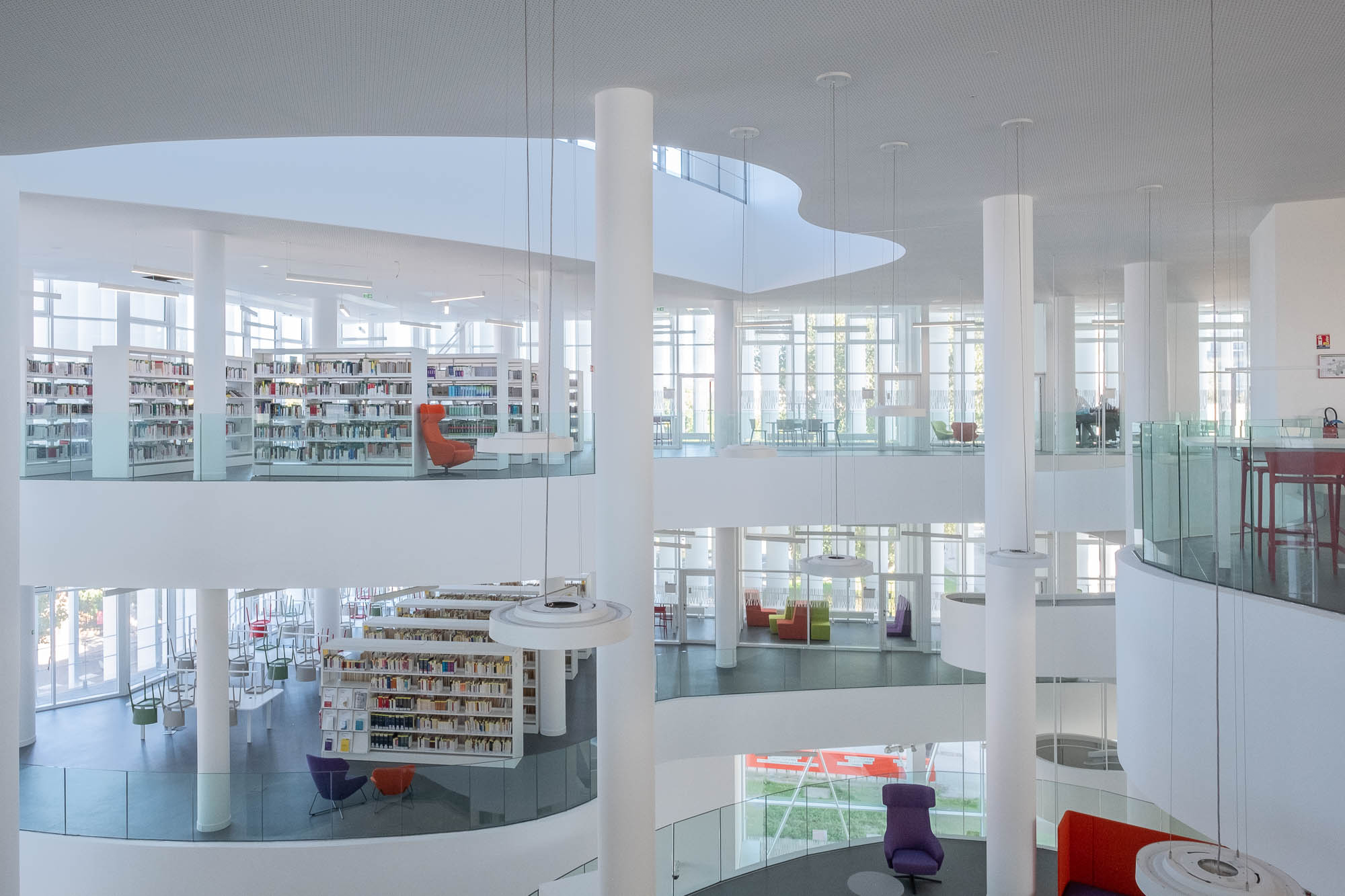 A contemporary, multilevel library with curved balconies and tall white columns. Filled bookshelves and colourful seats contrast against the grey and white surroundings. Natural light comes from large windows at the back.