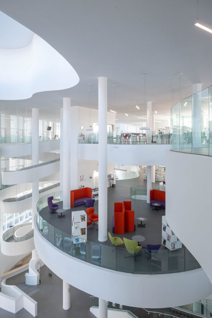 A contemporary, multilevel interior with curved balconies and tall white columns. Colourful seats contrast against the grey and white surroundings. Glass railings overlook an open area lit by an overhead light well.