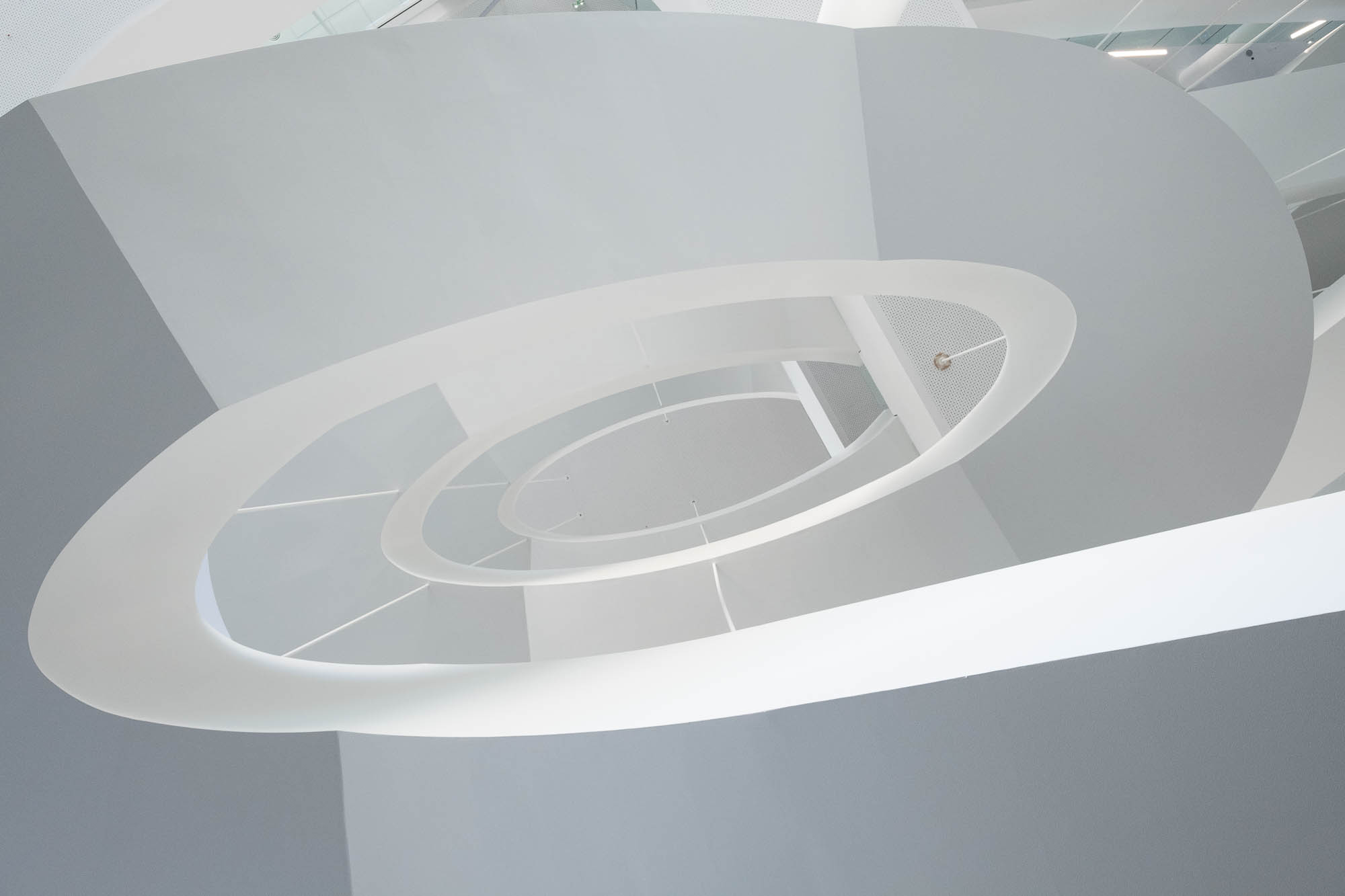 A contemporary architectural spiral staircase is viewed from below, showcasing its smooth, white curves and open circular design set against a bright background.